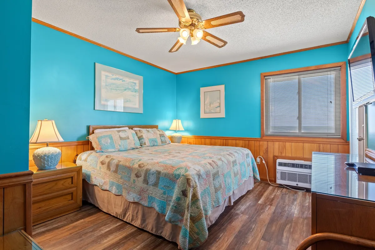 The image displays a cozy bedroom featuring a bed with colorful bedding, a nightstand with a lamp, a window with blinds letting in soft natural light, and artwork on the walls. The overall atmosphere is peaceful and inviting, emphasizing both comfort and style.