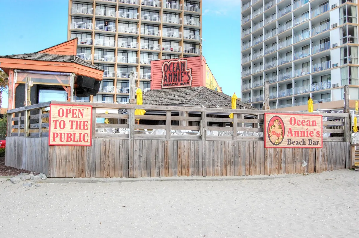 This image focuses on the iconic Ocean Annie's Beach Club sign, cheerful yellow umbrellas, and a wooden fence, all set against a lively, welcoming backdrop. There’s a sense of social energy with implied live music and drinks, echoing the beachside entertainment atmosphere.