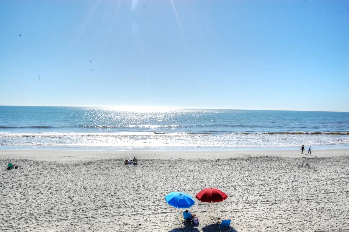 A lively stretch of sandy beach with blue and red umbrellas, families and children enjoying the sun, and sparkling ocean waves in the background. The atmosphere is playful, relaxed, and ideal for building sandcastles or wading in the water together.