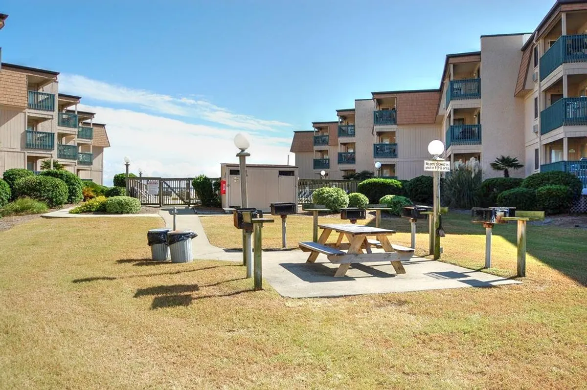 An exterior gathering space with picnic tables, grill stations, and landscaped greenery. Guests can be seen barbecuing, picnicking, and enjoying outdoor meals together. Balconies of adjacent buildings hint at easy access from accommodations.