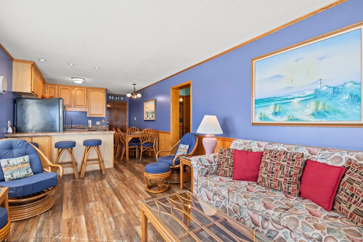 This interior shot features a colorful couch with decorative pillows, a wooden dining table and chairs, a kitchen counter with bar stools, and beach-themed artwork on the wall. The arrangement highlights a seamless flow between living, dining, and kitchen areas, evoking a cozy, social environment.