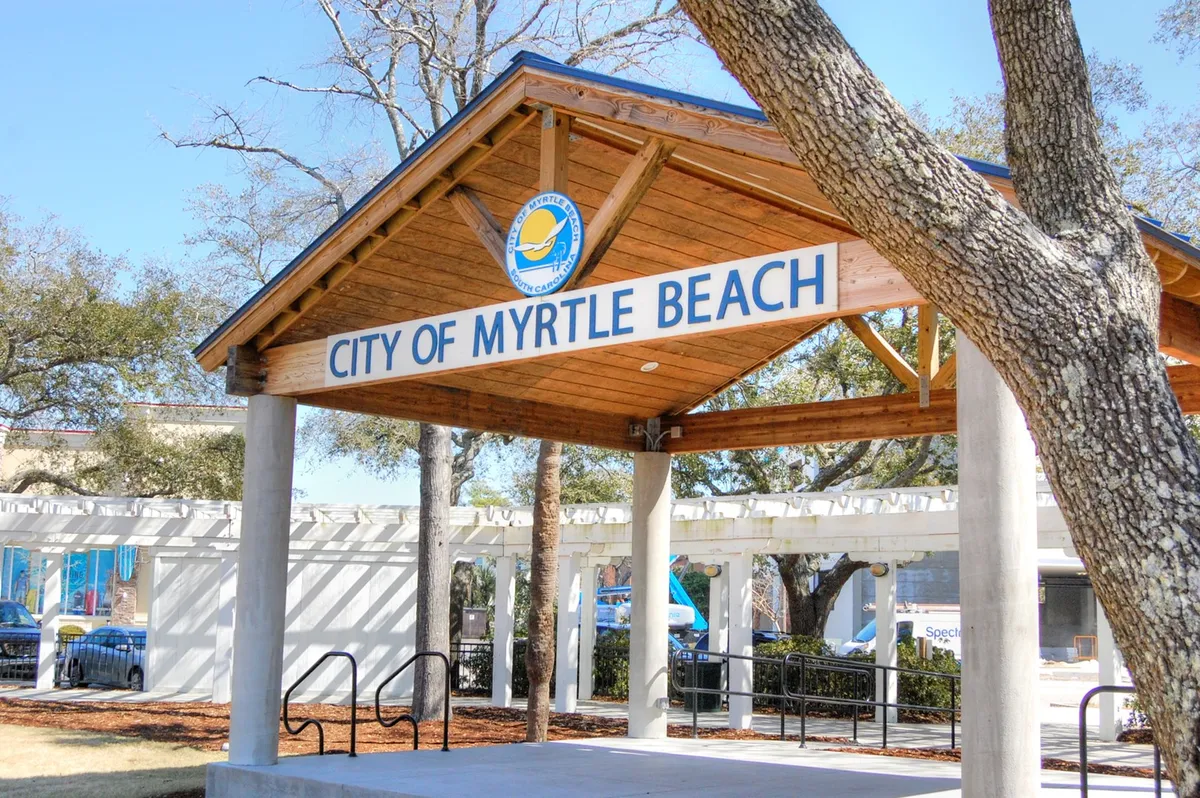 The welcoming entrance to Myrtle Beach featuring the iconic city sign, a charming wooden pavilion, and lush surrounding trees. The setting hints at community events and a friendly, vibrant atmosphere characteristic of the area.