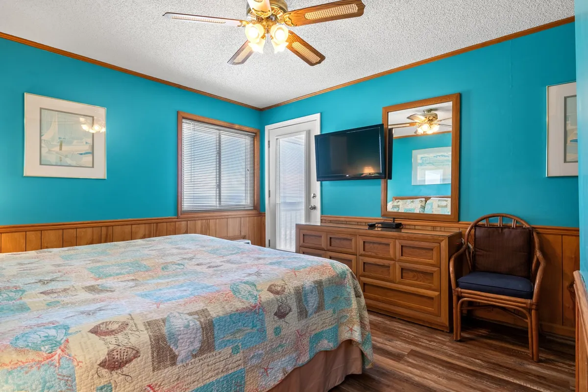 A cozy bedroom scene with a bed dressed in a colorful quilt, a mirror reflecting sunlight, and a wooden dresser. The room is designed for comfort, offering a sense of warmth and relaxation suitable for restful nights.