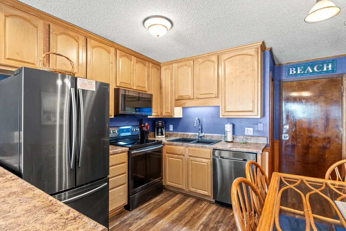 This interior view showcases the kitchen, featuring a black refrigerator, stainless steel appliances, wooden cabinets, and a playful beach-themed sign. The layout shows off the workspace and modern amenities ideal for preparing fresh meals.