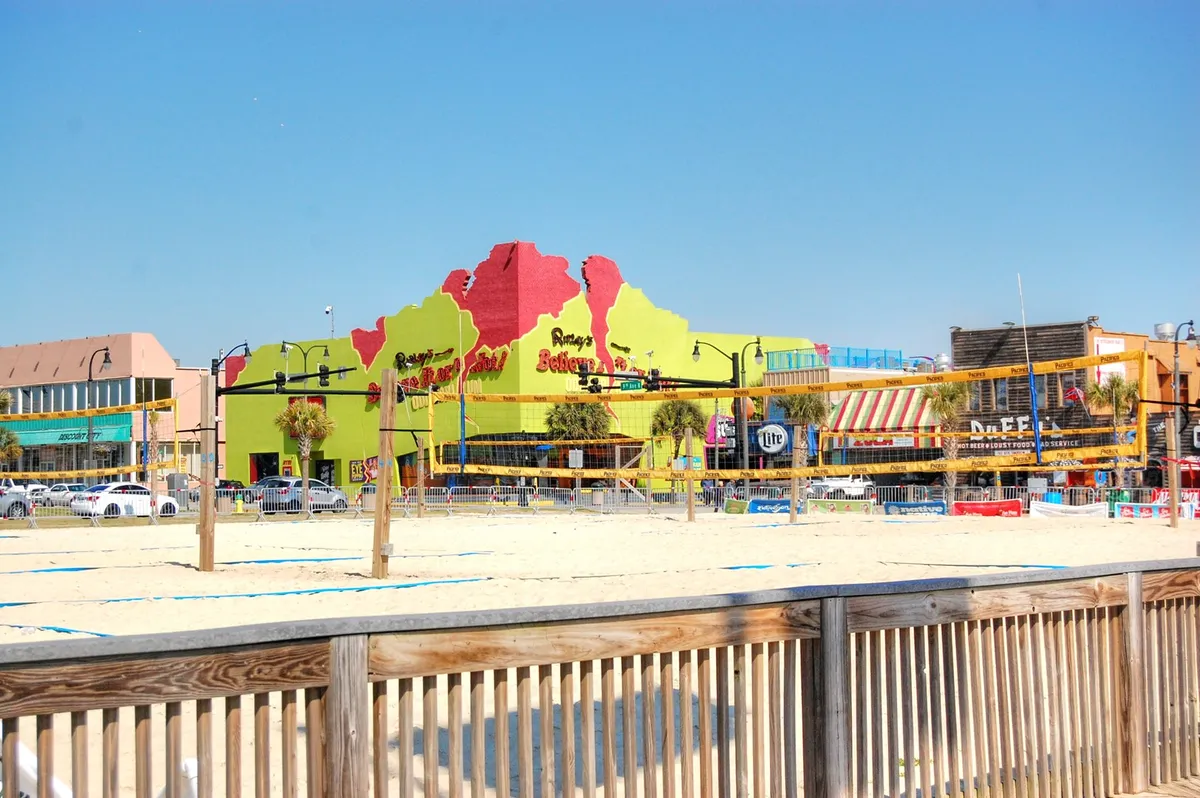 A colorful beachfront building facade sets the stage, with beach volleyball courts and a wooden boardwalk bustling with casual, energetic socializing. The scene hints at beach parties, group activities, and easy access to dining and fun.