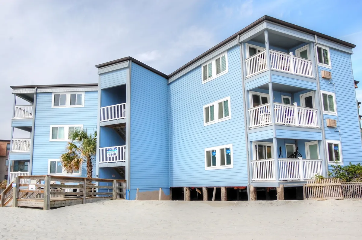 The blue exterior of a beachfront building with inviting balconies overlooking the sand, where families can gather to watch the sunset or enjoy evening breezes. The sandy ground below suggests immediate proximity to the beach.