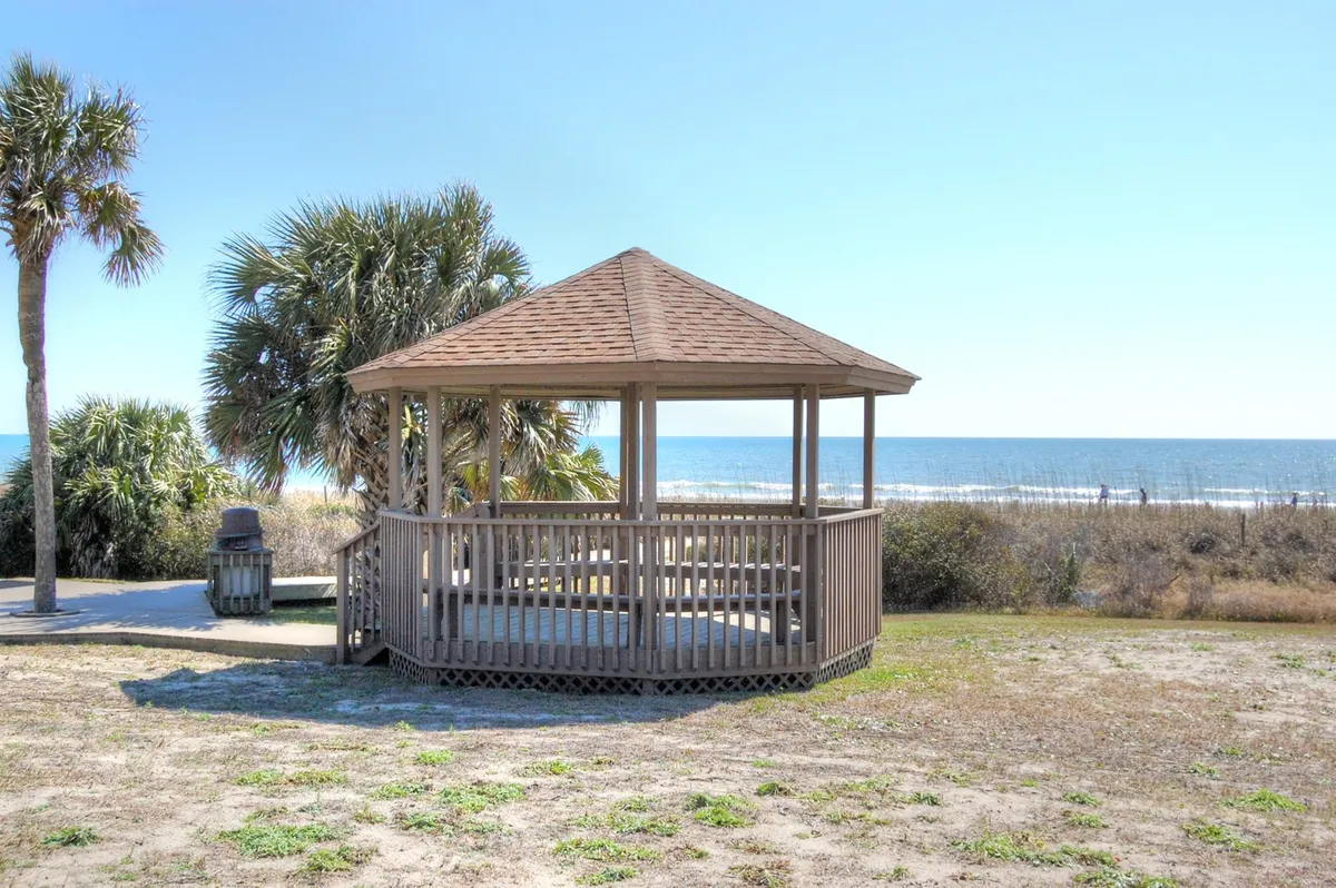 A serene exterior view featuring a charming gazebo overlooking the ocean, framed by palm trees and a tranquil shoreline. The setting is inviting for morning coffee or sunset relaxation with the sound of the waves in the background.