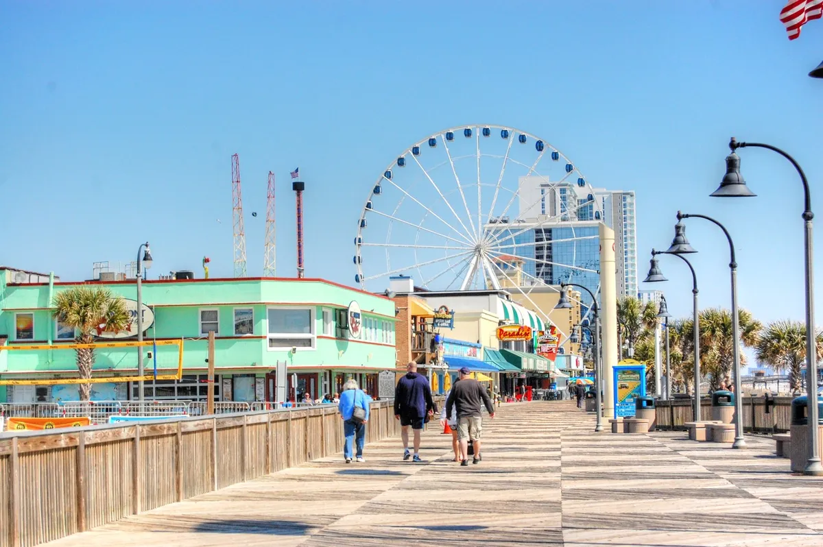A lively boardwalk scene featuring a colorful ferris wheel, palm trees, and vibrant buildings. The boardwalk is bustling with visitors enjoying rides, snacks, and the festive atmosphere—hinting at local attractions and entertainment options.