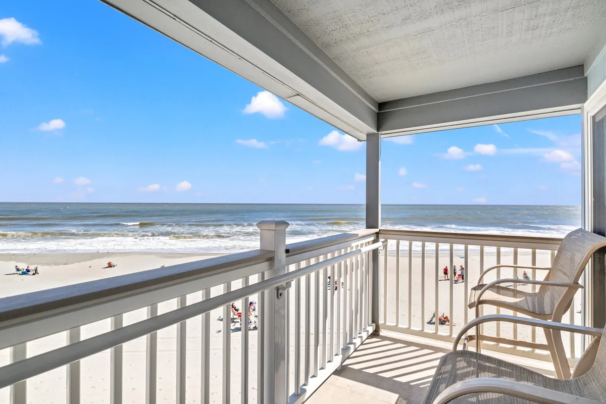 The image shows an inviting balcony overlooking the wide expanse of Myrtle Beach, with comfortable seating perfectly positioned to soak in panoramic views of the ocean, gentle waves rolling ashore, and the vibrant atmosphere of Ocean Annie’s nearby. The scene is tranquil, with golden sunlight illuminating the sand and surf, suggesting both sunrise coffee moments and sunset dinners.
