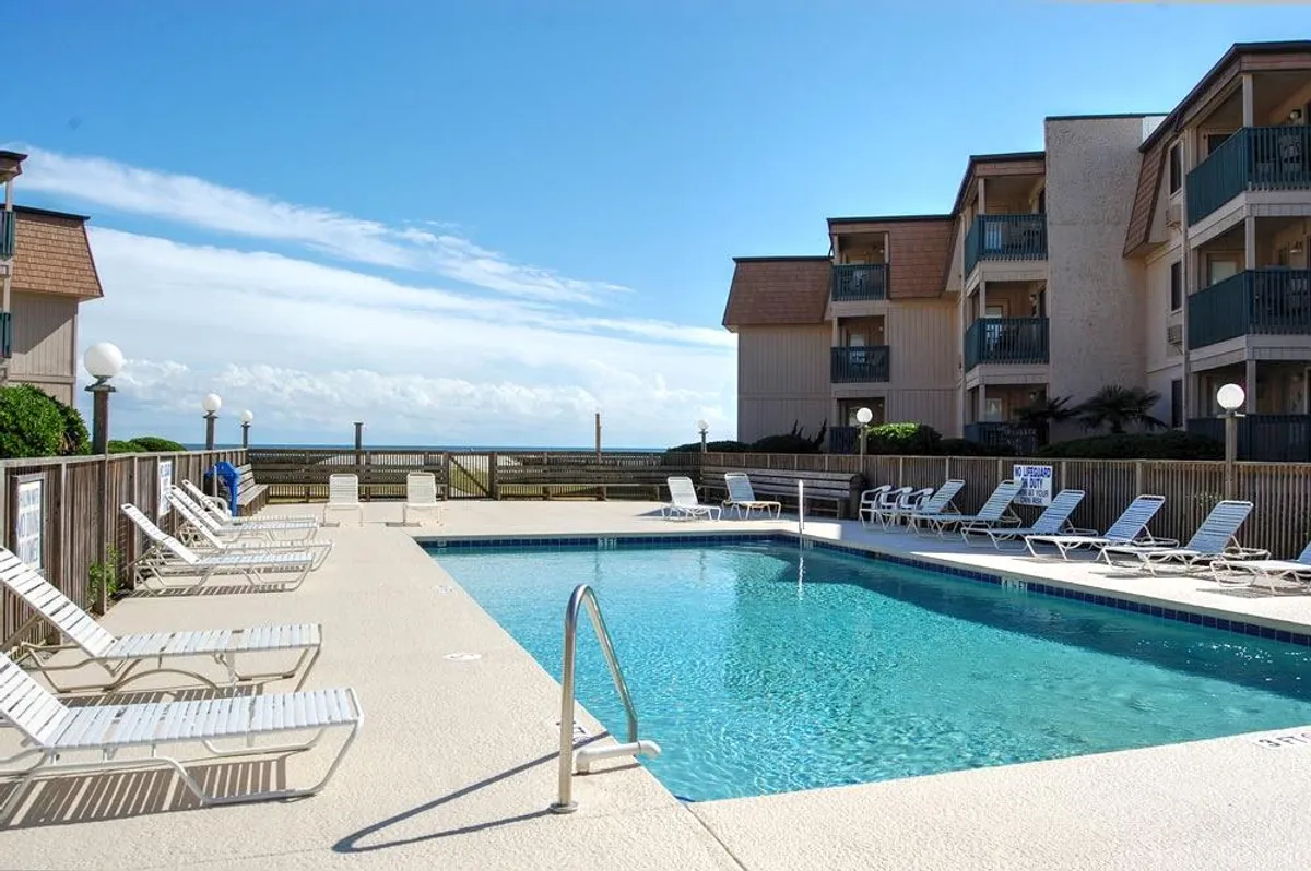 A sparkling swimming pool surrounded by lounge chairs and overlooking the ocean. The building's balconies are visible in the background, inviting guests to relax and socialize poolside. The atmosphere is cheerful and relaxed, ideal for vacationers seeking leisure and fun.