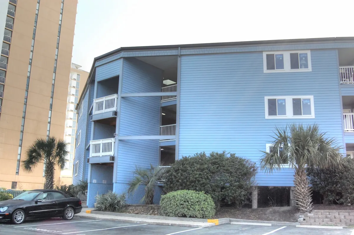 An inviting exterior shot of the blue resort building, flanked by palm trees and with balconies visible, suggesting private spaces for enjoying fresh air and ocean views. The mood is casual yet welcoming, hinting at both resort amenities and personal comfort.