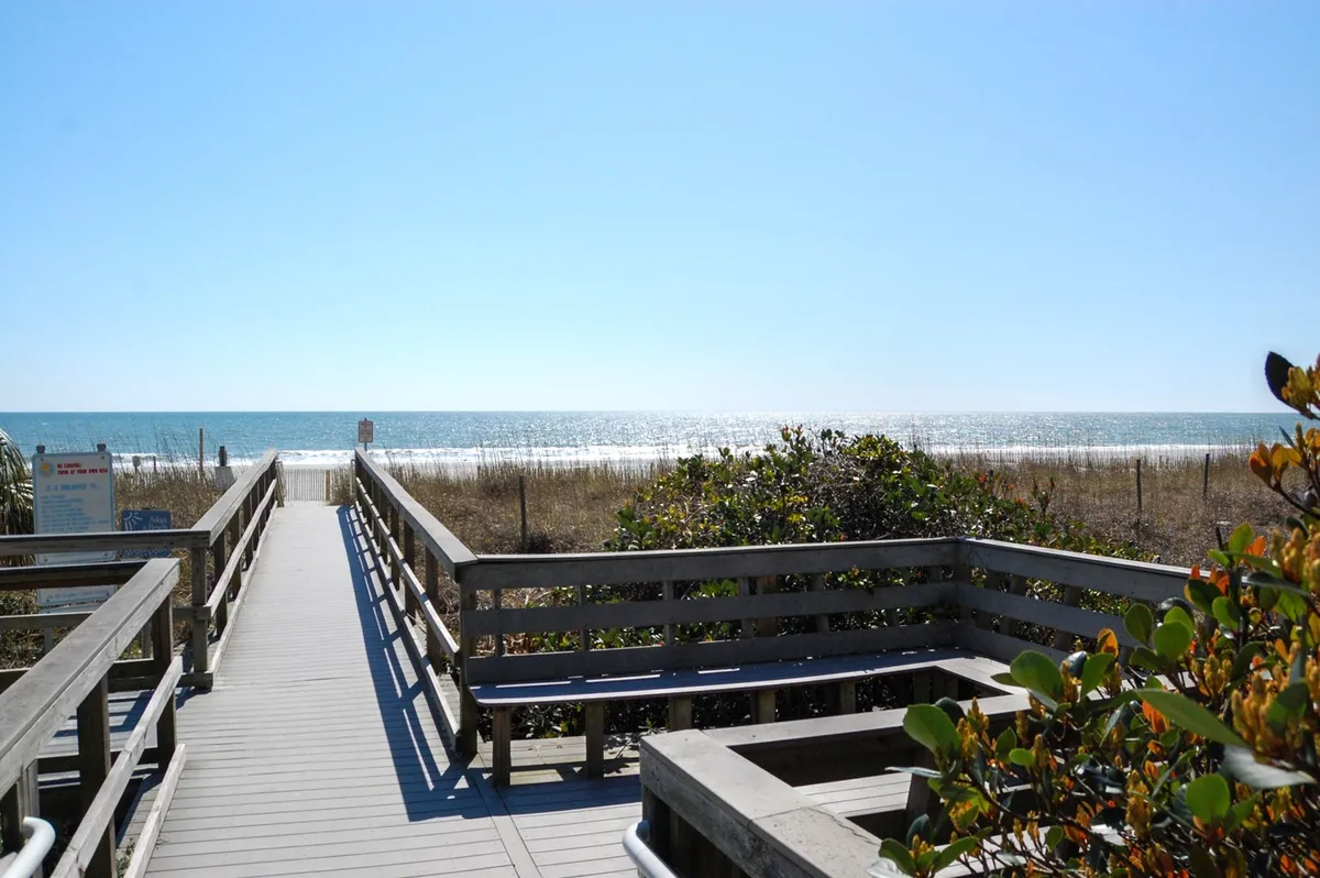A serene view along a wooden walkway leading through beach grass toward the open ocean and a distant horizon. The scene is tranquil, inviting guests to imagine peaceful strolls to the water's edge or relaxing moments by the beach, enhanced by soft natural light.