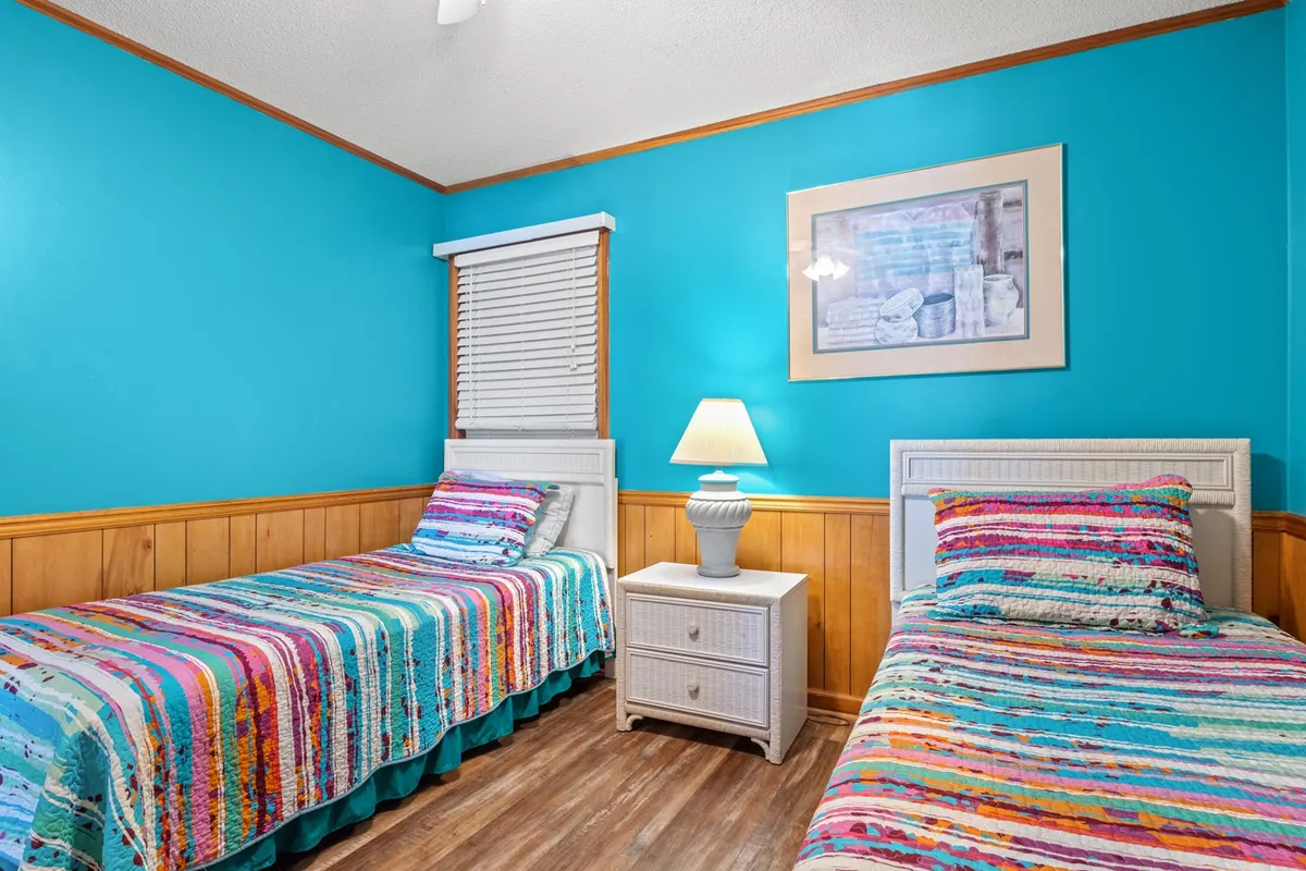 This bedroom features colorful striped bed covers, white furniture, blue wall art, and a table lamp. The playful yet relaxing décor creates a welcoming space ideal for children or friends. The room is well-lit and arranged for both comfort and fun.