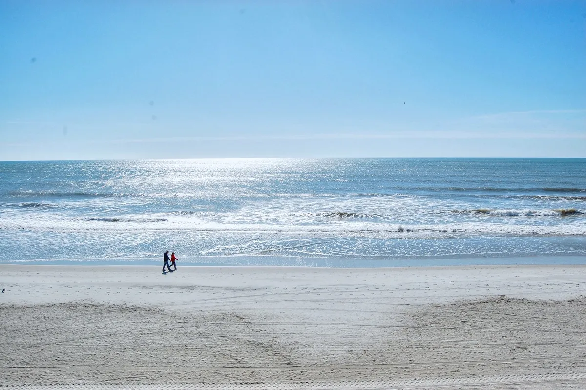 A romantic scene featuring a couple walking hand-in-hand along a sunlit beach, with gentle ocean waves rolling in and a bright, expansive sky overhead. The focus is on the couple enjoying a peaceful, intimate moment by the water, framed by the serene beachfront landscape.