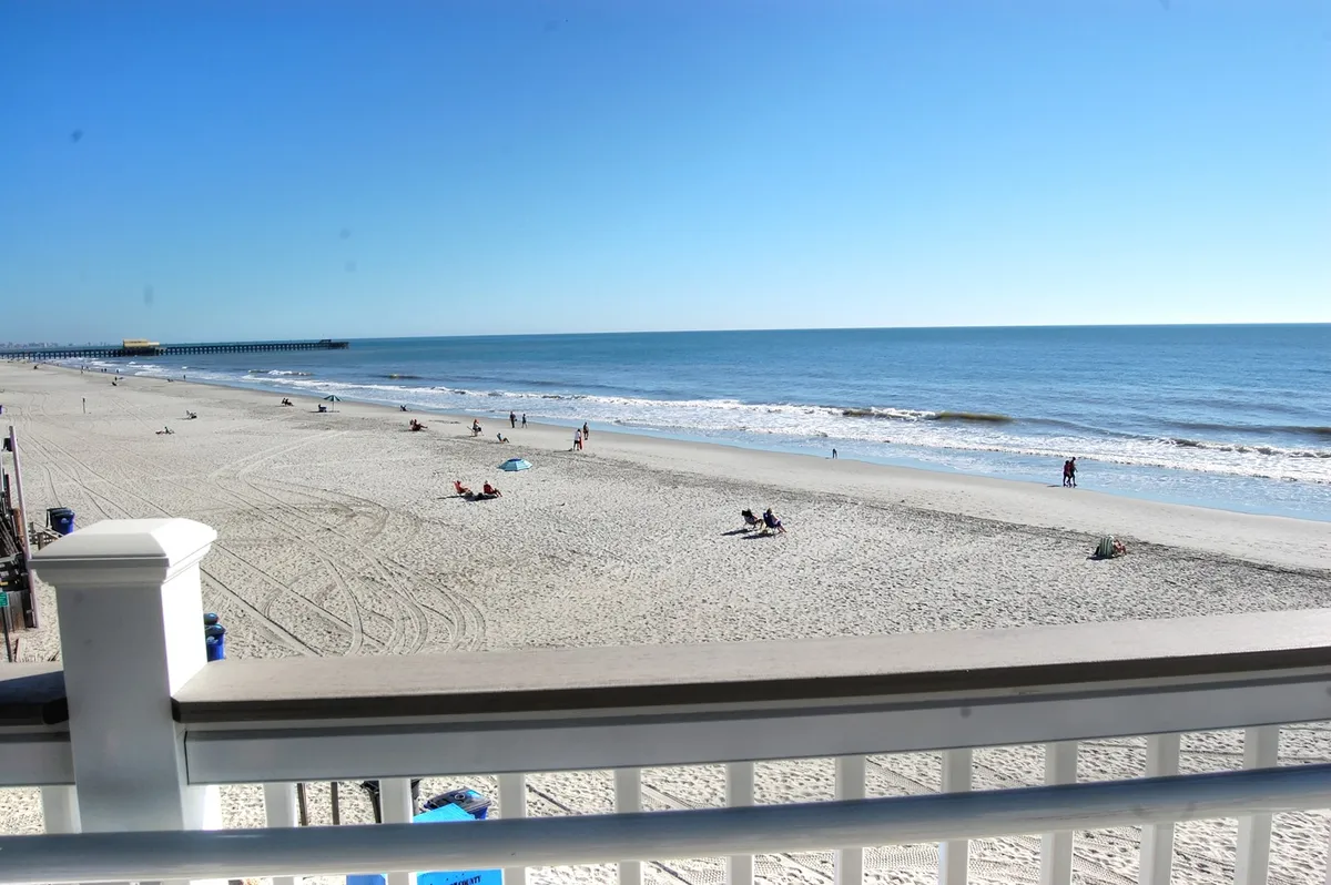 A vibrant beachfront scene with people sunbathing, swimming, walking along the shore, and enjoying the sunny weather. The pier stretches into the ocean, providing a lively and inviting atmosphere.