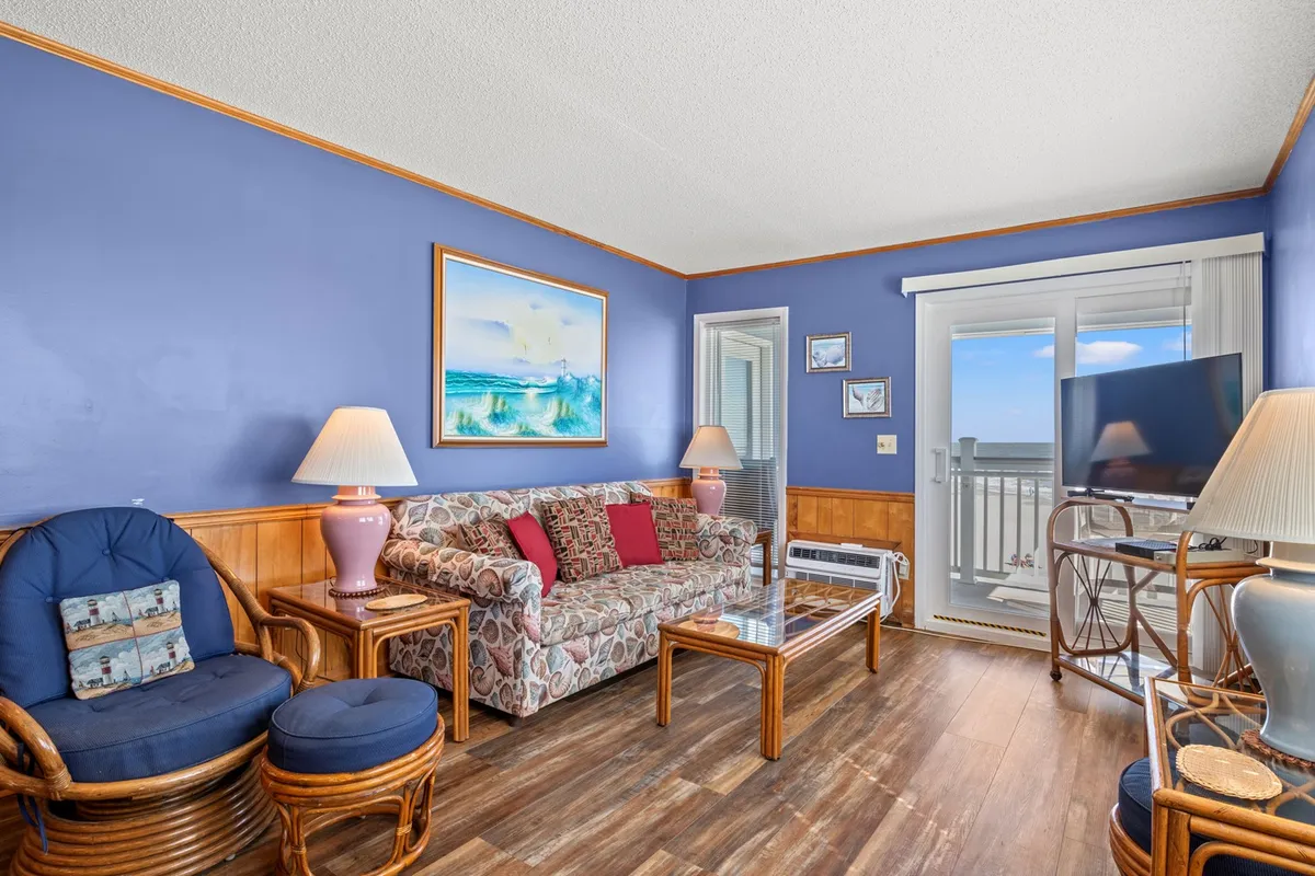 A cozy living room with a comfortable sofa positioned to take in the ocean view, colorful artwork adorning the walls, and warm lighting from decorative lamps. Sliding glass doors open to a balcony, inviting natural light and ocean breezes into the space. The scene radiates comfort, ideal for relaxing, unwinding with a movie, or socializing with loved ones.